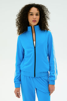 Wearing the SPLITS59 Fox Techflex Jacket in Riviera/White, a person with curly hair shows off retro style in matching blue and white activewear, standing against a simple light background.