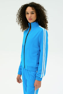 A person with curly hair wears the SPLITS59 Fox Techflex Jacket in Riviera/White, featuring a retro vibe and white sleeve stripes, standing against a plain white background.