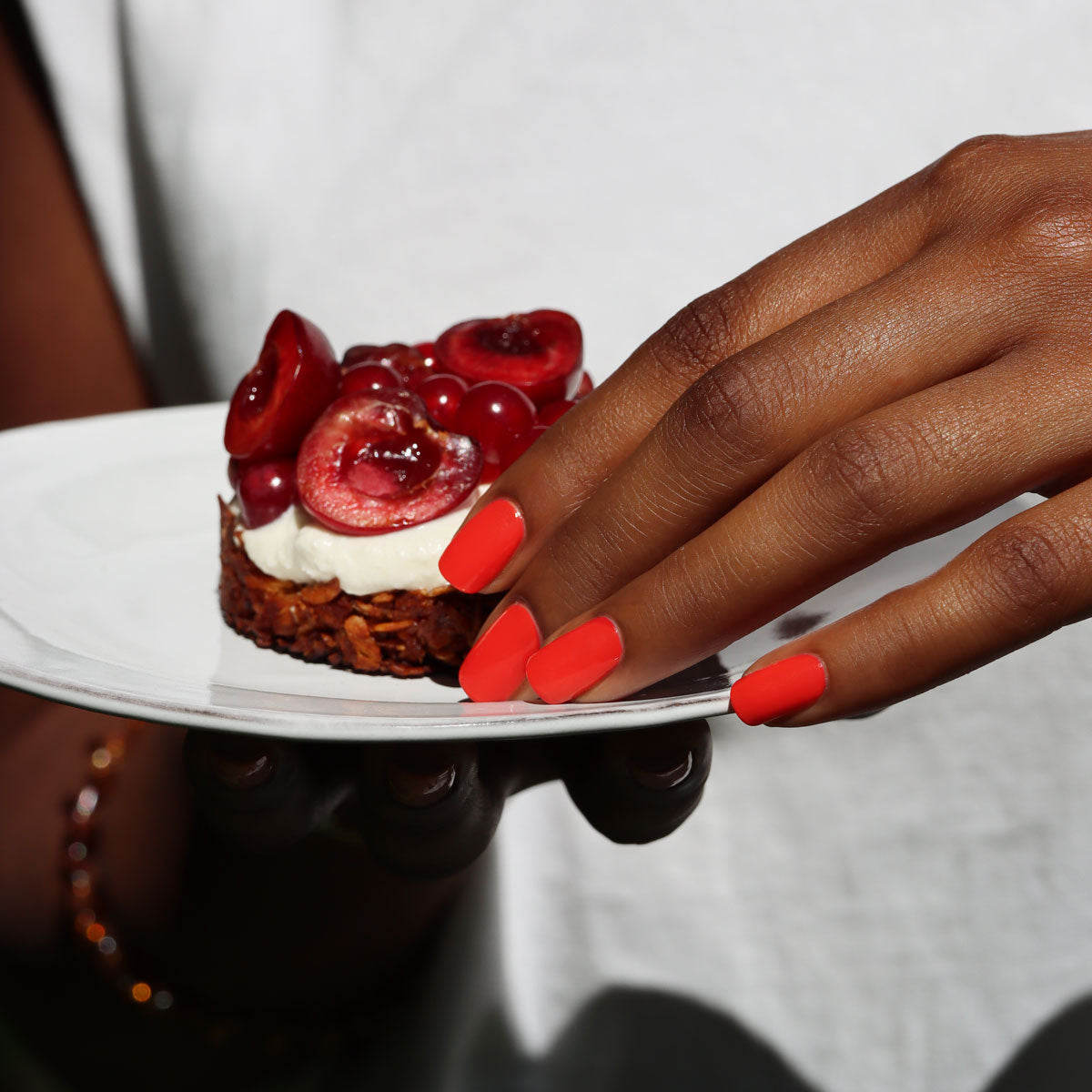 A person with bright orange nail polish holds a white plate with a small dessert topped with cream and fresh cherries. The person is wearing a white shirt.
