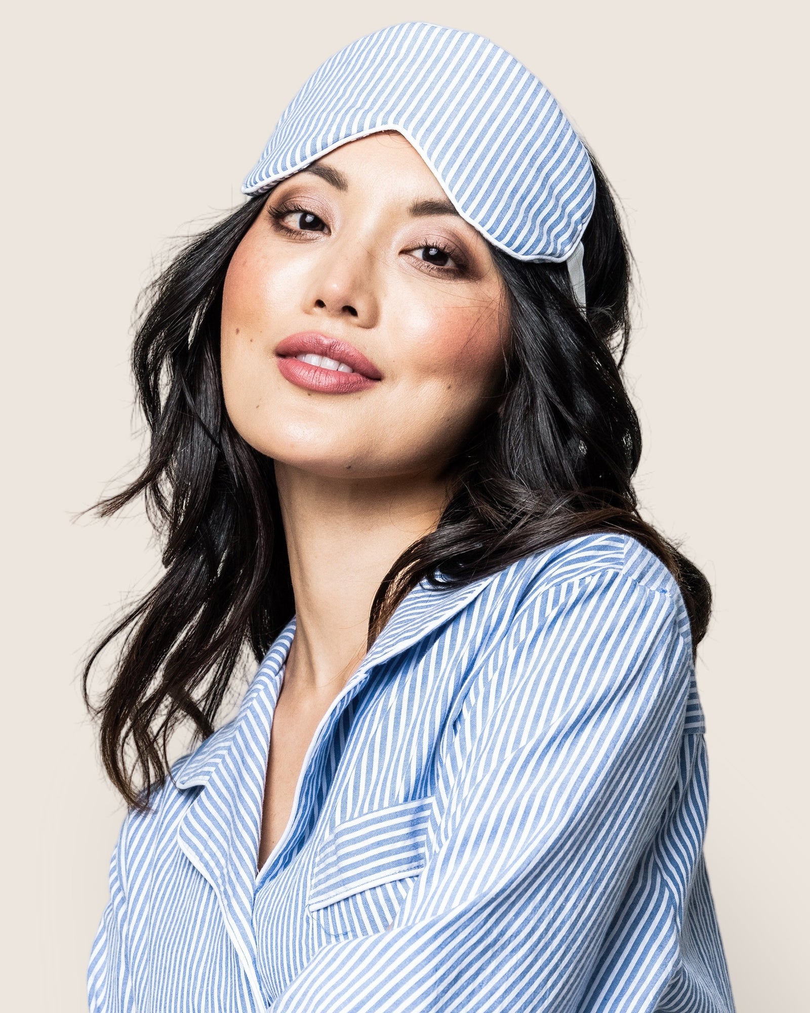 An individual with long dark hair wears Petite Plumes Adults Sleep Mask in French Blue Seersucker atop their head, complemented by matching blue and white striped pajamas. Their neutral expression exudes luxury sleep against a light background.