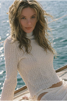 Woman wearing a cream-colored textured long-sleeve top with a turtleneck, posed on a boat with water in the background.
