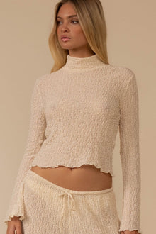 Cream textured long-sleeve top with mock neckline and scalloped hem, shown on model against neutral background.