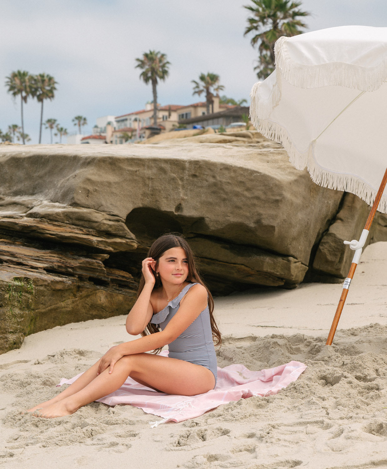Young girl wearing a striped one piece bikini sitting on a towel on the beach.