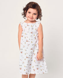A young girl with short brown hair smiles in the Girl's Twill Amelie Nightgown in Easter Frolic, a white sleeveless dress featuring playful animal and nature prints, standing against a plain light background.