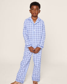 A young boy sports Petite Plumes Kids Twill Pajama Set in Royal Blue Gingham. He is barefoot with short, curly hair, and a plain background highlights his luxury sleepwear’s gingham print.