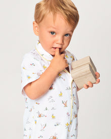 A young child with blonde hair wears the Baby's Twill Summer Romper in Easter Frolic—white with colorful animal prints and yellow trim—standing against a plain light background, holding two wooden blocks and looking to the side.