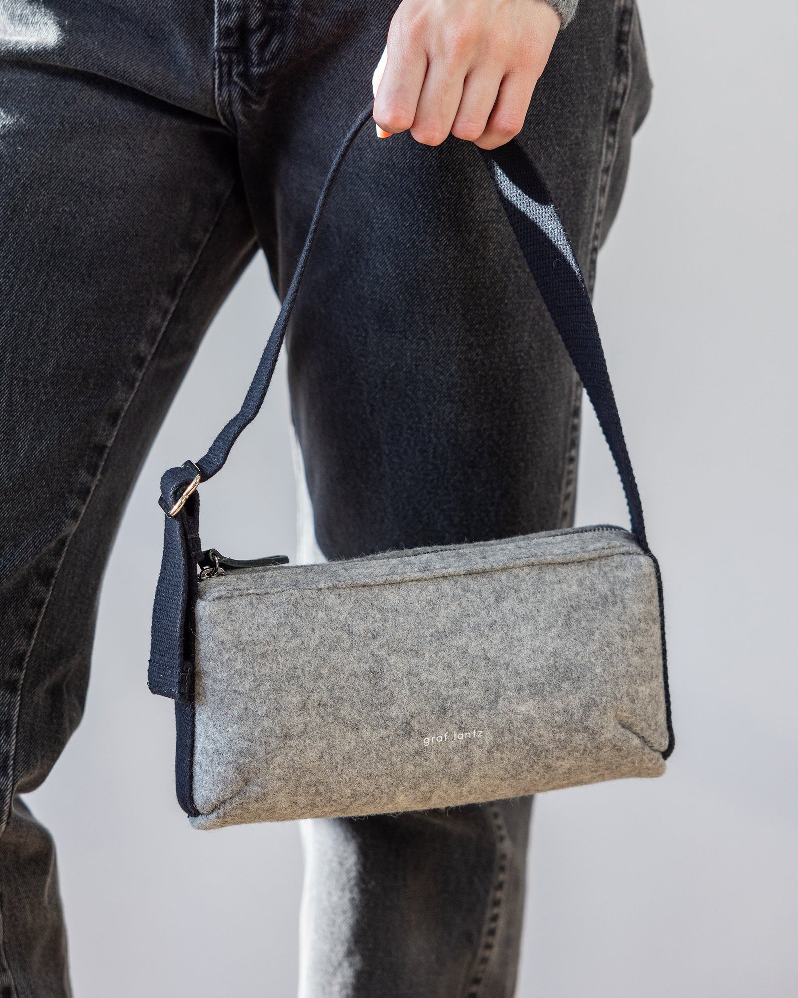 A person holds a small, gray felt graf lantz Campus Shoulder Bag with a black strap. Ideal for everyday essentials, the bag complements their dark jeans against the plain light gray background.