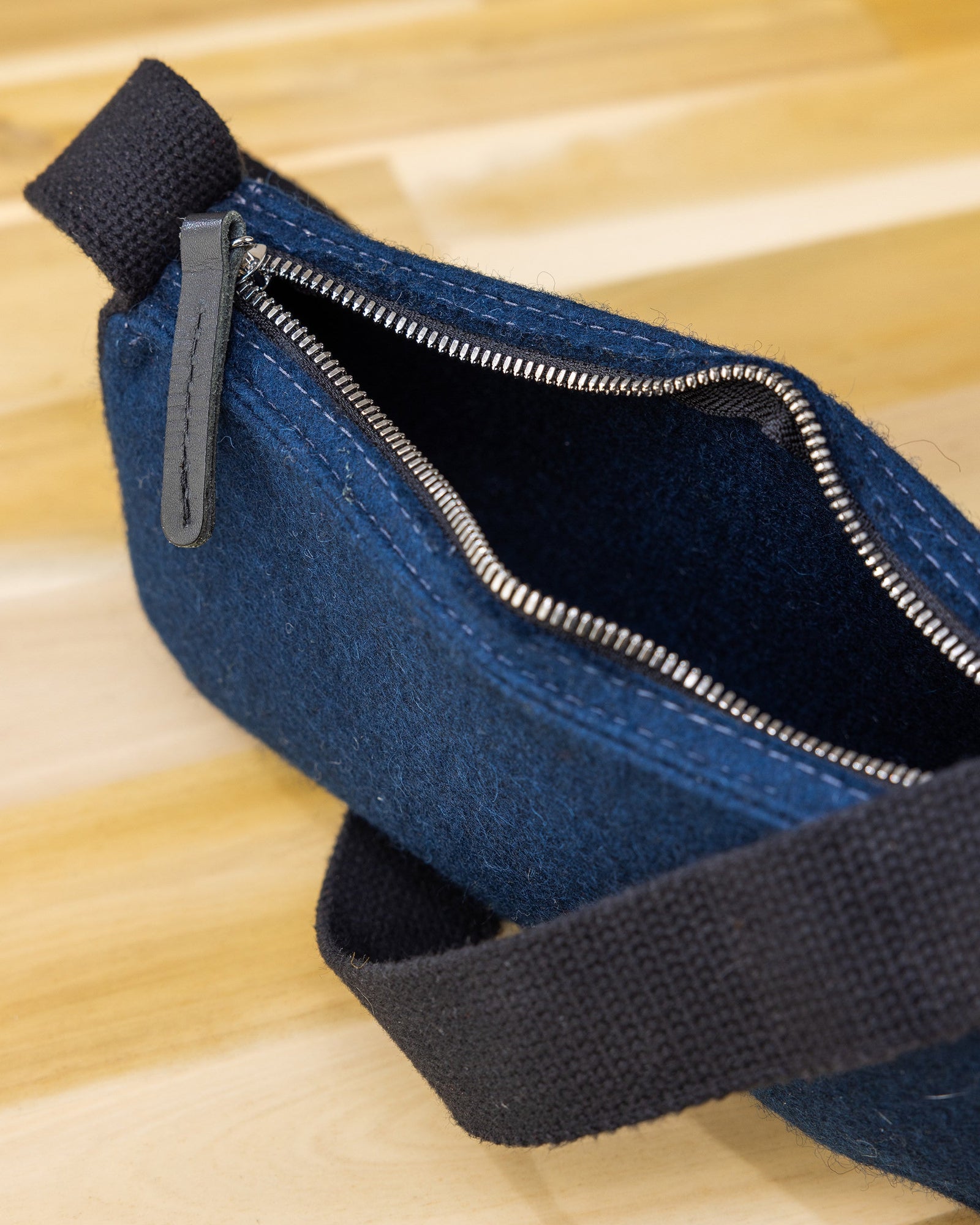 A close-up of a graf lantz dark blue felt pouch showcases its minimalist design with a partially open zipper and black fabric loop handle on wood. Its texture, similar to Merino Wool, appears soft as the metallic zipper glides smoothly along the top edge.