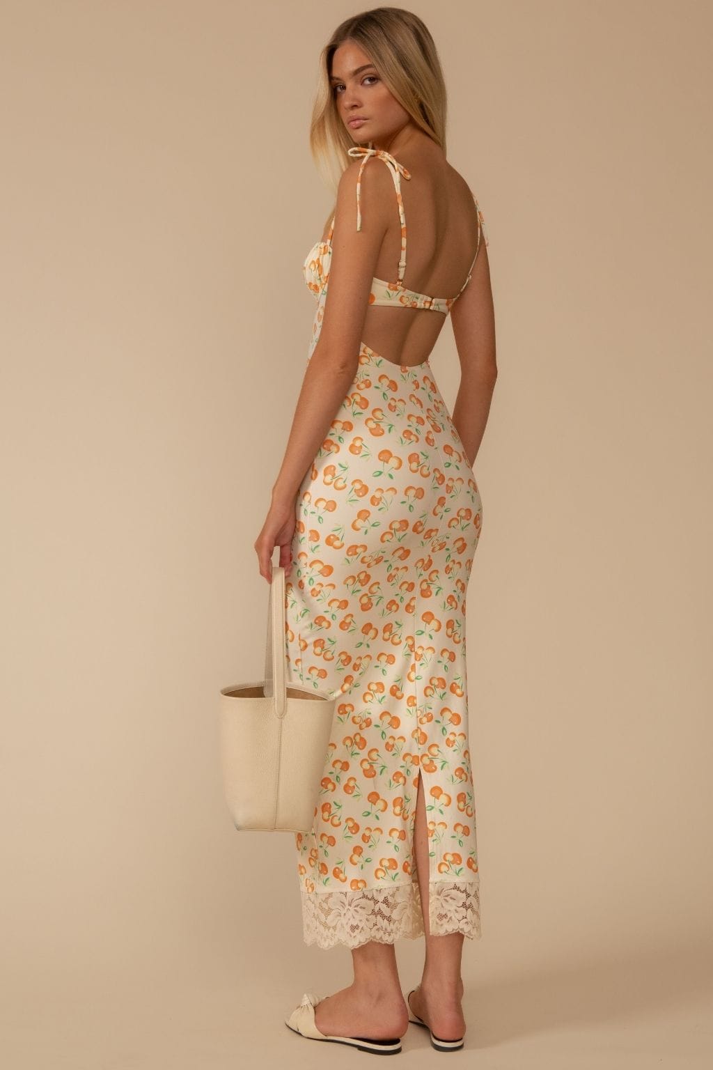 Woman wearing a long slip dress with cherry print, open back, and lace trim at hem, styled with a cream tote bag.