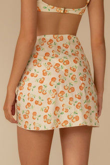 Cream sarong with all-over orange cherry and green leaf print, worn as a wrap skirt.