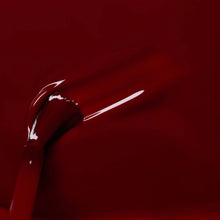 A close-up of a brush applying glossy dark red nail polish, with visible brush strokes and reflections on the smooth surface.