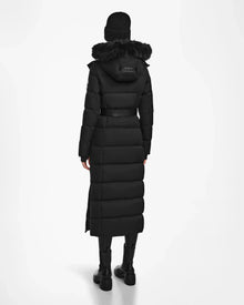 A person stands facing away against a plain white background, wearing the Rudsak IZABELLA - L124541 Black women's long down coat with a fur-trimmed hood, paired with a black knit hat and black boots.