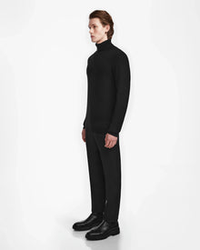 A person with light skin and straight, brown hair stands in profile against a white background, wearing the Rudsak NICOLAS - 6524528 Black mens cashmere turtleneck, along with black trousers and shoes.