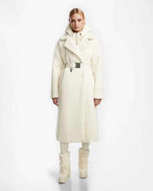 A woman faces forward in a long, white Rudsak TINA - 8123578 Iceland faux fur teddy trench coat with a wide collar and silver belt, styled with white pants and boots against a plain white background.