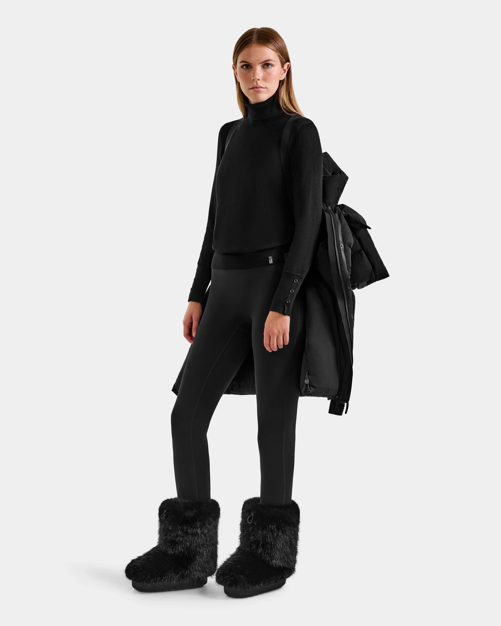 A woman faces forward against a light background, wearing the Rudsak ALESSANDRA M - L125552 Black winter coat draped over one shoulder, paired with a black turtleneck, black leggings, and fluffy black boots.