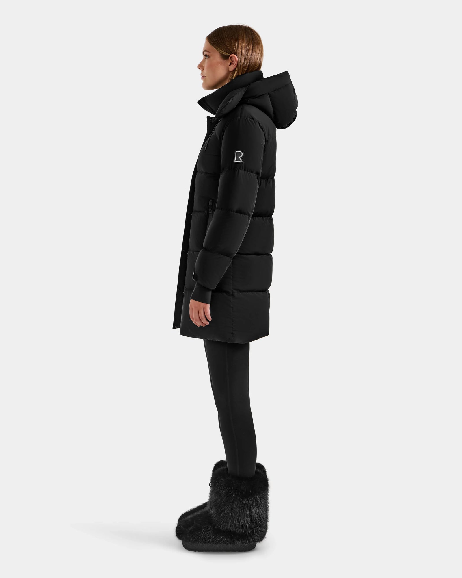 A person stands in profile against a plain light background wearing the Rudsak ALESSANDRA M - L125552 Black women’s down puffer with a hood, paired with black leggings and black furry boots.