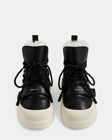 A pair of Rudsak ALEXEY L - L225518 Black women’s leather winter boots with thick white soles, black laces, and visible plush wool lining, shown from the front on a plain white background.