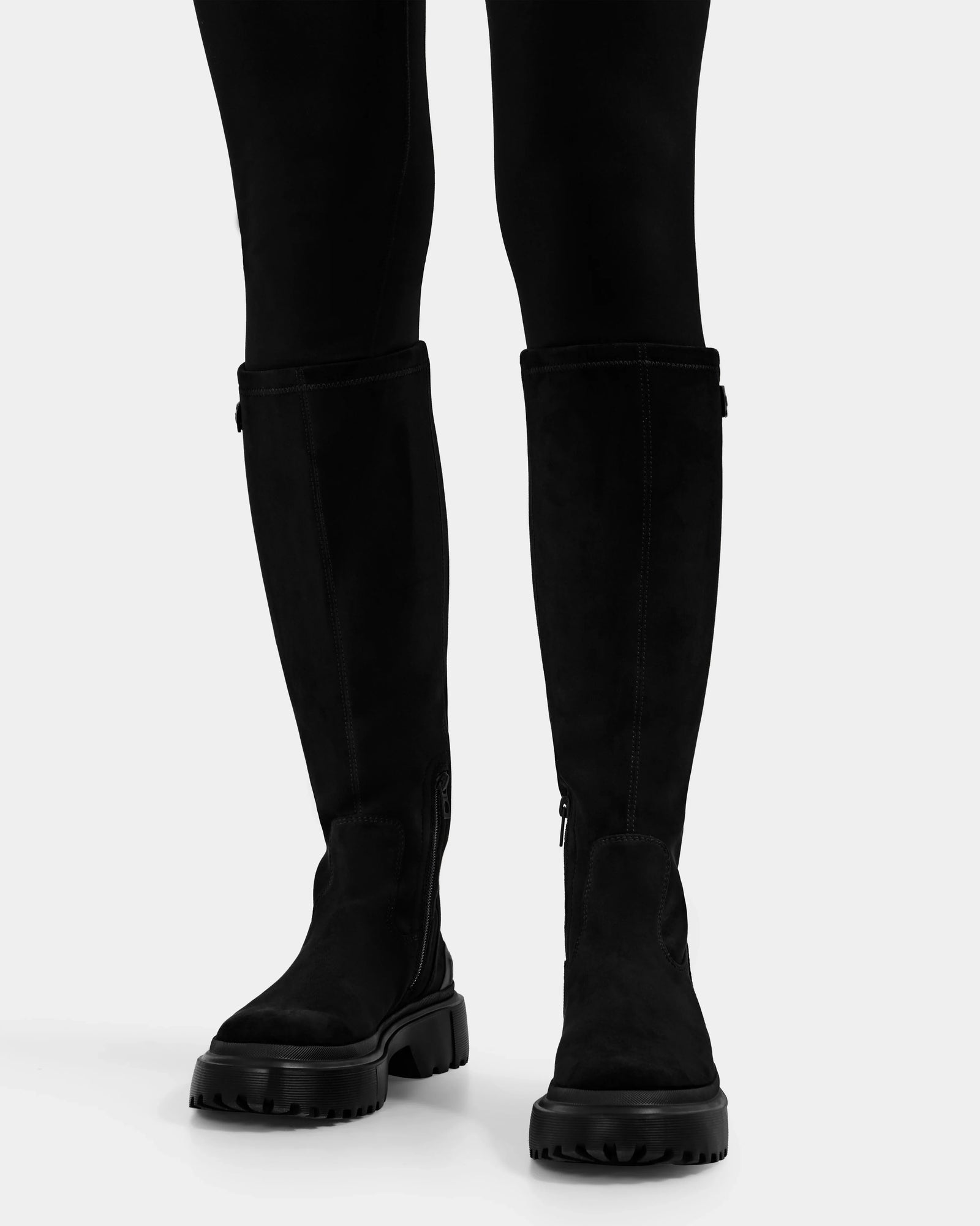 A person wearing Rudsak’s AVOLA - L225514 Black faux-suede knee-high boots with chunky soles and black leggings, standing on a white background.