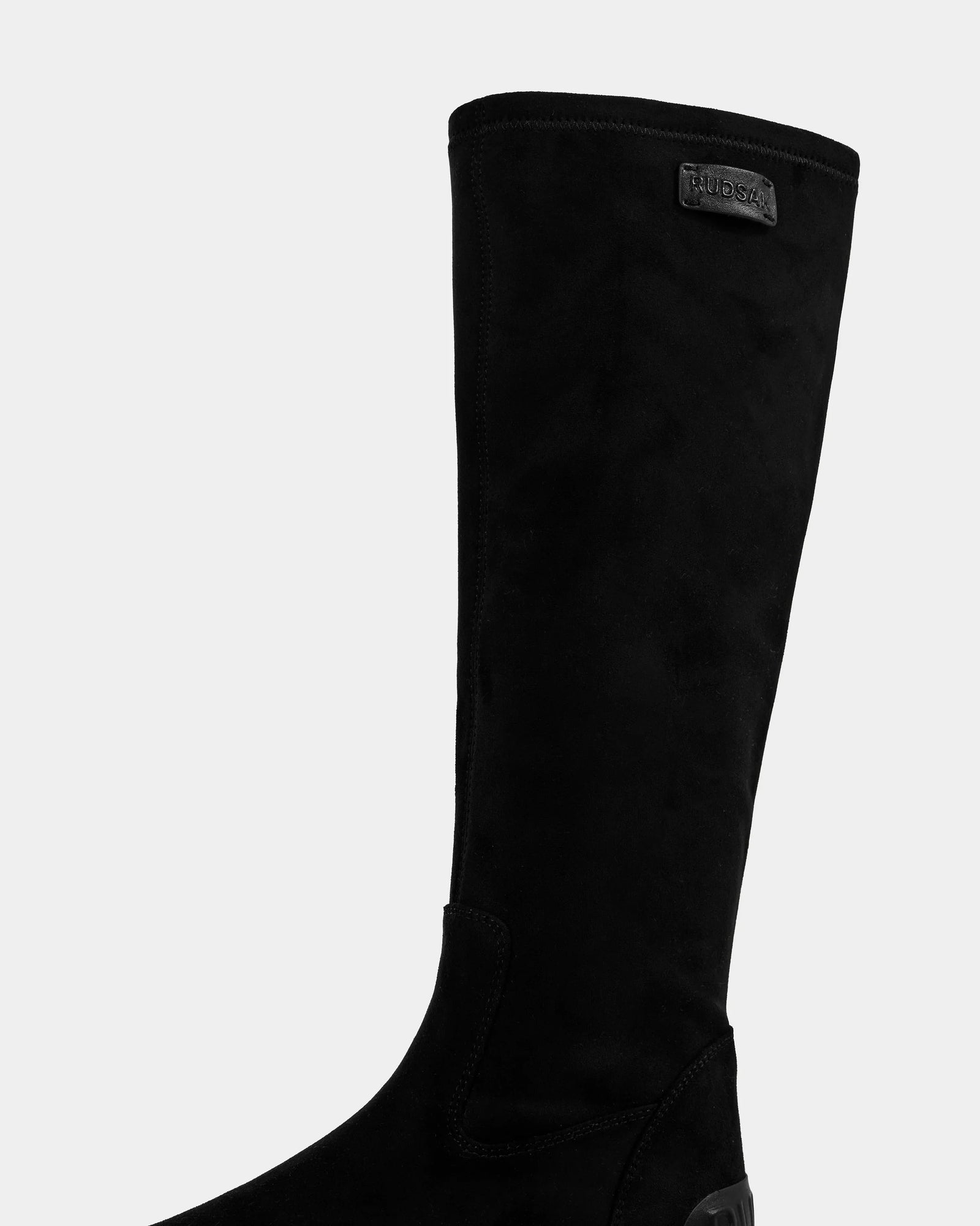 A close-up of the AVOLA - L225514 Black boot by Rudsak, featuring a tall black faux-suede shaft with a small rectangular logo patch near the top, set against a plain light background.