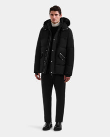 A man faces forward wearing the Rudsak BRADY - M125510 Black men’s quilted down parka with removable hood, paired with black pants, black shoes, and a white turtleneck against a plain light gray background.
