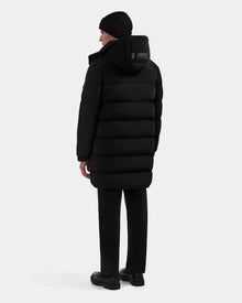 A person wearing the Rudsak BRAYDEN M-M - M125535 Black down puffer coat, black pants, shoes, and a beanie stands facing away against a plain light background, showcasing the coat’s winter versatility.