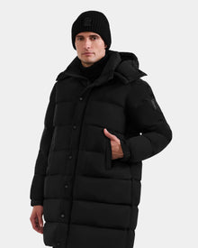 A person models the Rudsak BRAYDEN M-M - M125535 Black men’s down puffer coat with hood, paired with a black beanie, standing against a light gray background and highlighting its winter versatility.