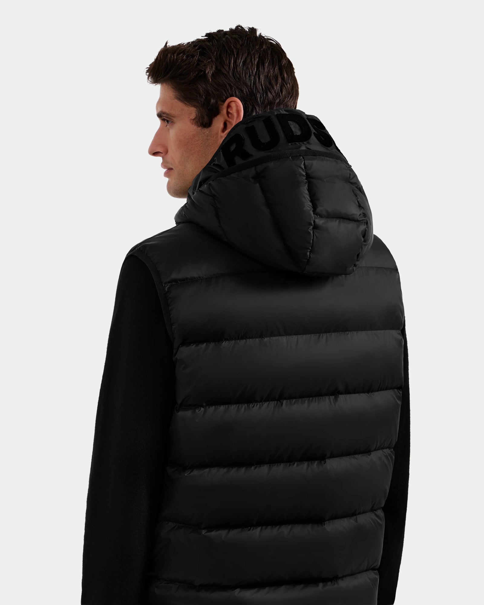 A man with short brown hair is seen from the back wearing the Rudsak BROOKS - M125520 Black puffer jacket, featuring a hood crafted from recycled-nylon shell with bold black lettering. The background is plain and light-colored.