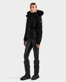 A woman stands against a light gray background wearing the Rudsak CHRISTELLE M - L125542 Black weather-resistant jacket with a fur-trimmed hood, paired with black leggings and knee-high boots.