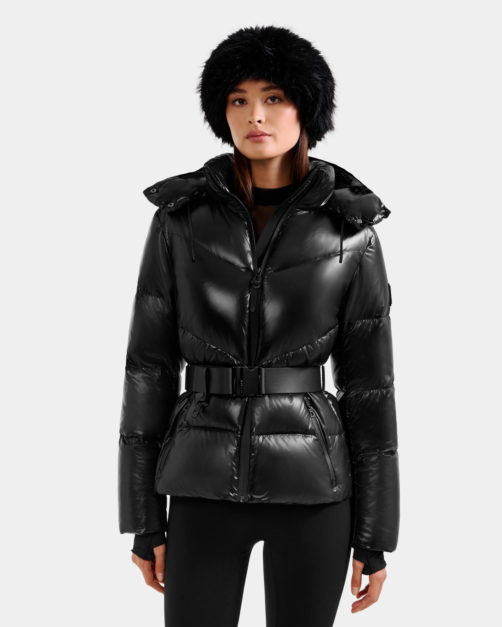 A woman in the Rudsak CHRISTELLE S - L125544 Black shiny winter jacket with a belt, black leggings, and a black furry hat stands against a plain light gray background.