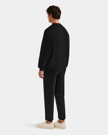 A man stands facing away, wearing the Rudsak COLSON - M525501 Black crewneck sweatshirt with matching black pants and white sneakers, set against a plain light gray background.