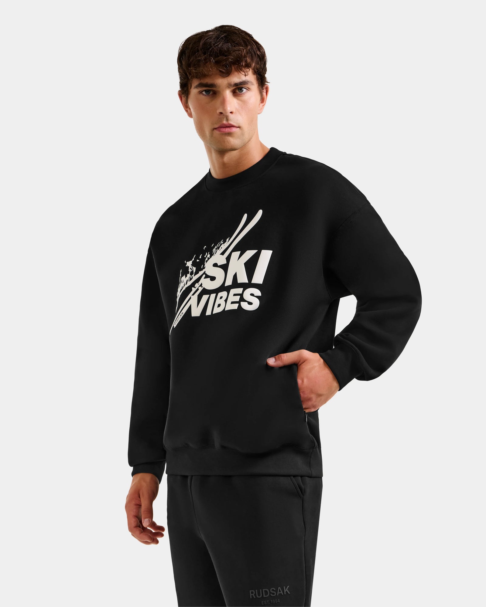 A young man in the Rudsak COLSON - M525501 Black men’s crewneck sweatshirt with SKI VIBES logo and ski graphics stands against a plain background, one hand in his pocket, exuding effortless après-ski style as he looks at the camera.