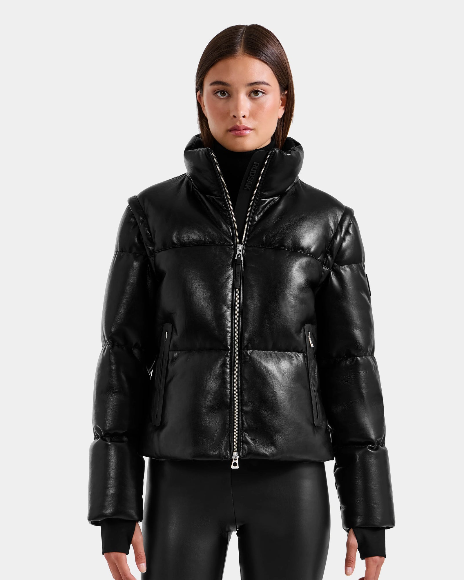 A woman with straight brown hair wears the Rudsak DELILAH PX - L125537 Black vegan-leather puffer with removable sleeves and black fitted pants, standing against a plain light gray background, looking neutrally at the camera.