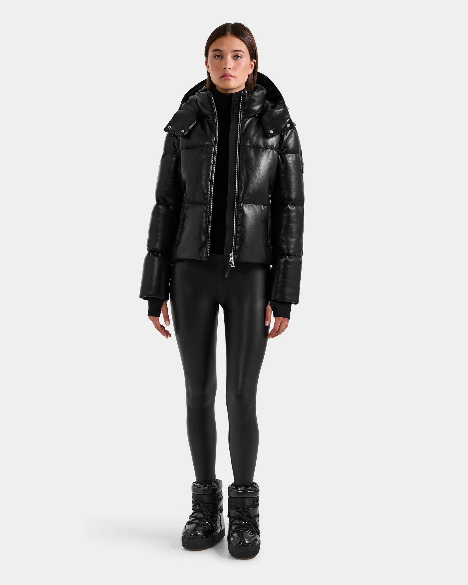 A person faces forward against a plain white background, wearing the Rudsak DELILAH PX - L125537 Black women’s vegan-leather puffer jacket, paired with black leggings and black shoes.