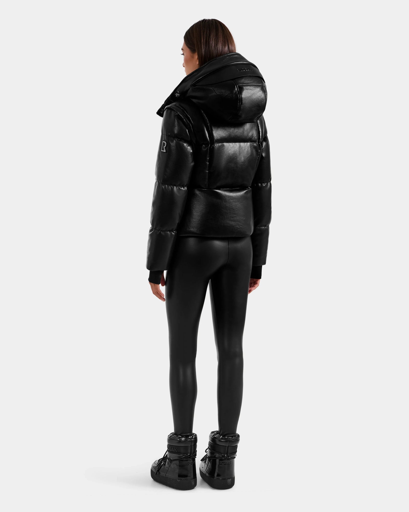 A person stands facing away, wearing the Rudsak DELILAH PX - L125537 Black vegan-leather puffer jacket, black fitted leggings, and black high-top shoes against a plain light gray background.