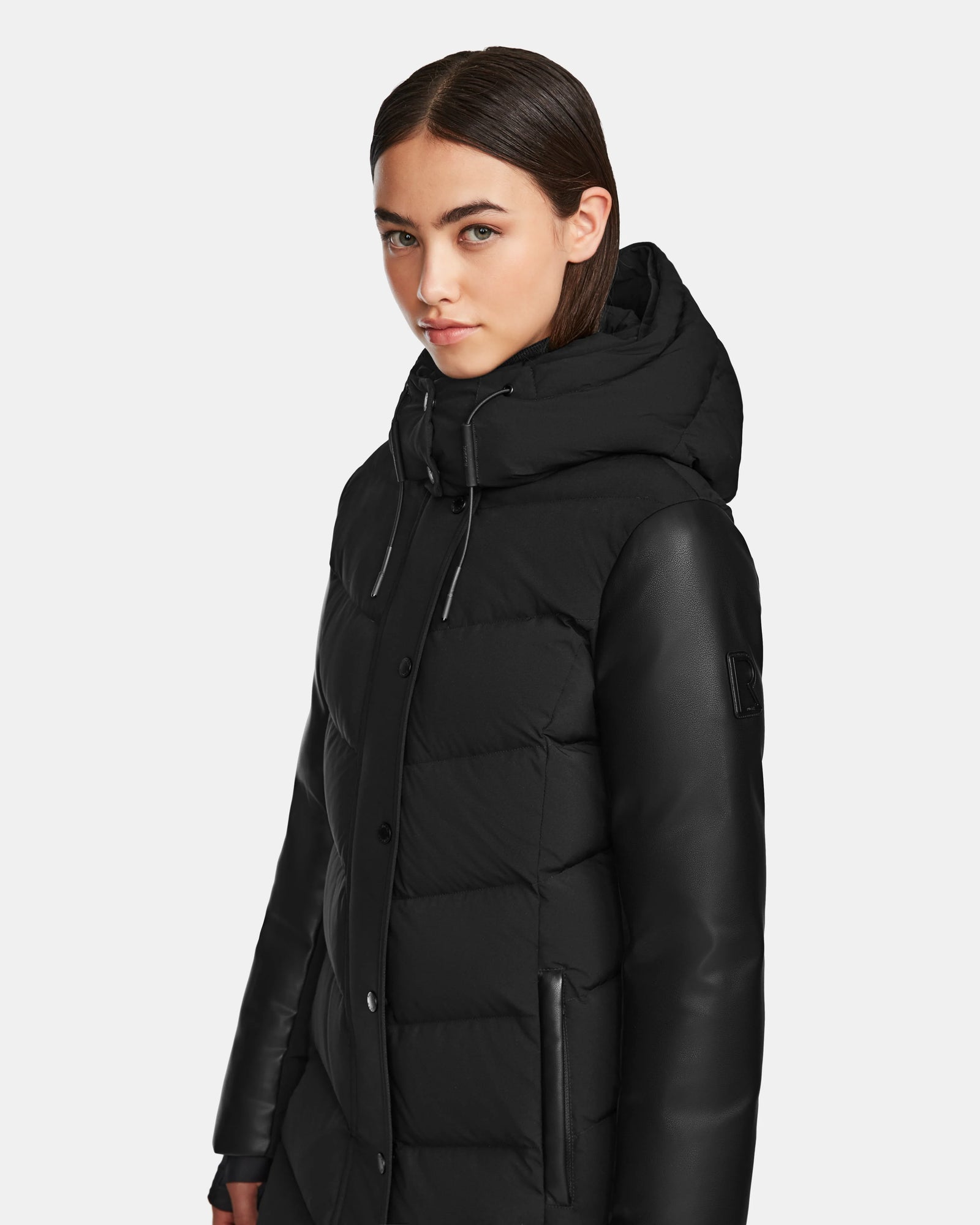 A woman with straight brown hair wears the Rudsak ELSA - L123535 Black sustainable winter jacket with a hood and leather sleeves, looking slightly to the side against a plain background.