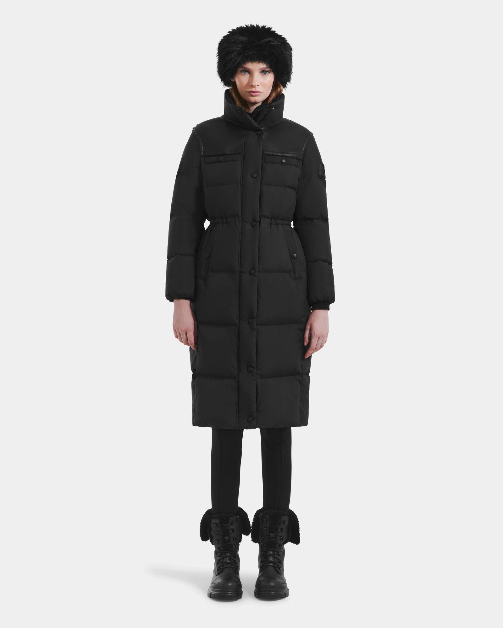 A person wearing the Rudsak EMBER M-L - L125543 Black women's long down puffer coat with a removable hood stands facing forward against a plain white background.