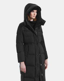 A woman stands slightly left against a white background, wearing the Rudsak EMBER M-L - L125543 Black women's long down puffer coat with a large removable hood and snap buttons.