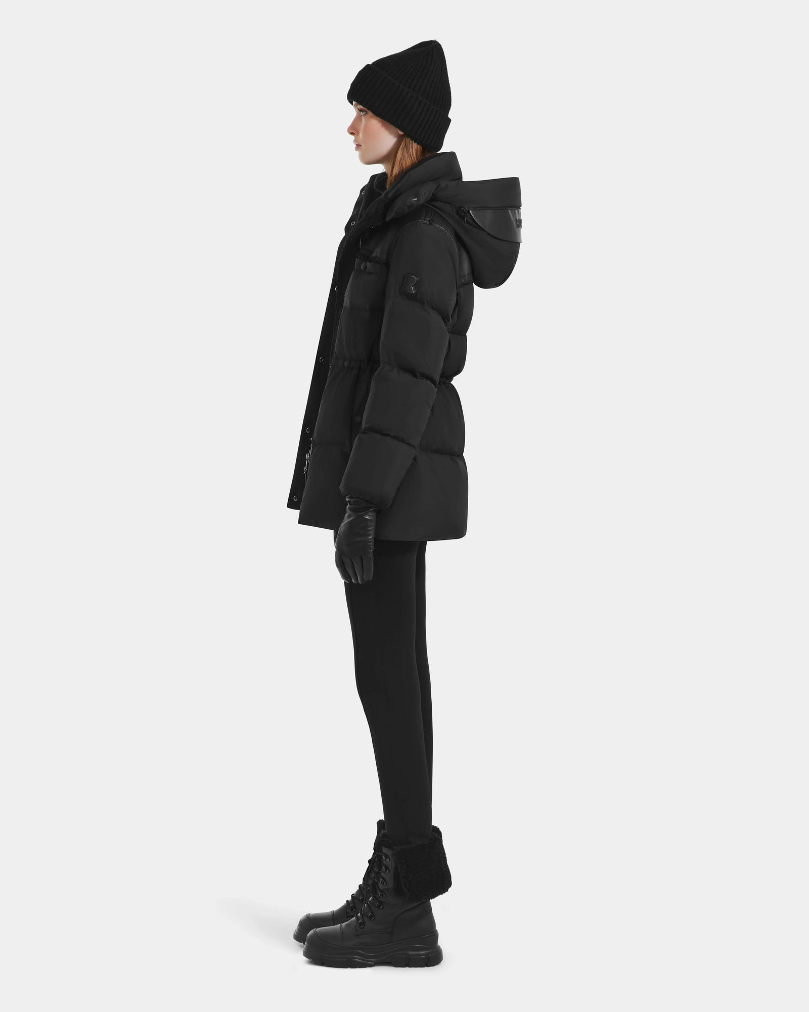 A person in profile wears the Rudsak EMBER M-M - L125524 Black women's down puffer with vegan-leather piping, black beanie, gloves, leggings, and lace-up boots, standing against a plain white background.