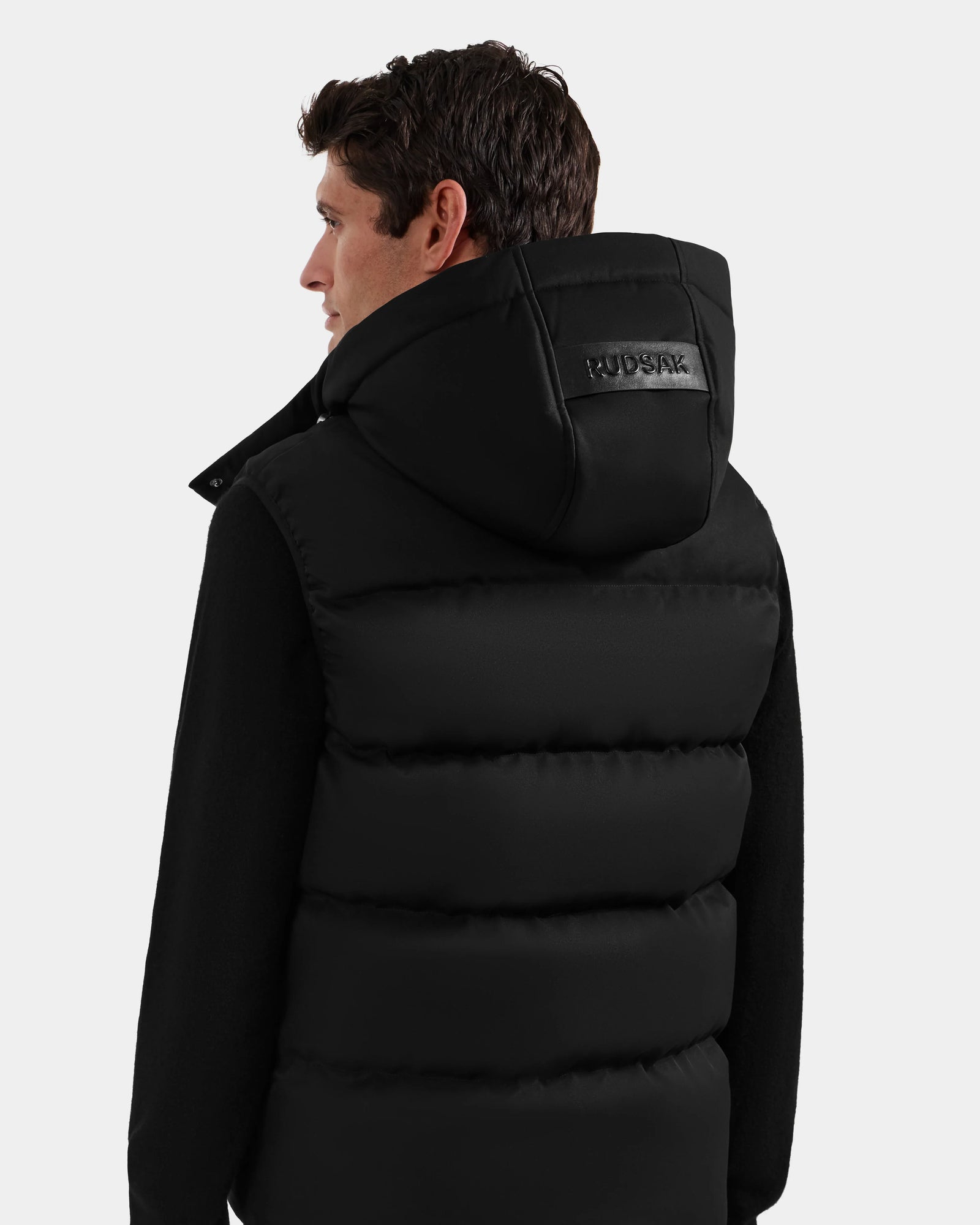A man with short brown hair is seen from the back wearing the Rudsak HARLOW - M125528 Black men’s puffer vest with "RUDSAK" on the removable hood, over a black long-sleeve top, against a plain light background.
