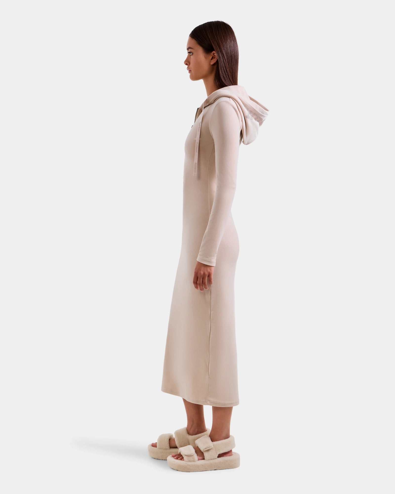 A woman in profile wears the Rudsak ISABELLA - L525502 Cream velour hooded dress with long sleeves and fuzzy sandals, featuring a subtle logo, set against a plain white background.