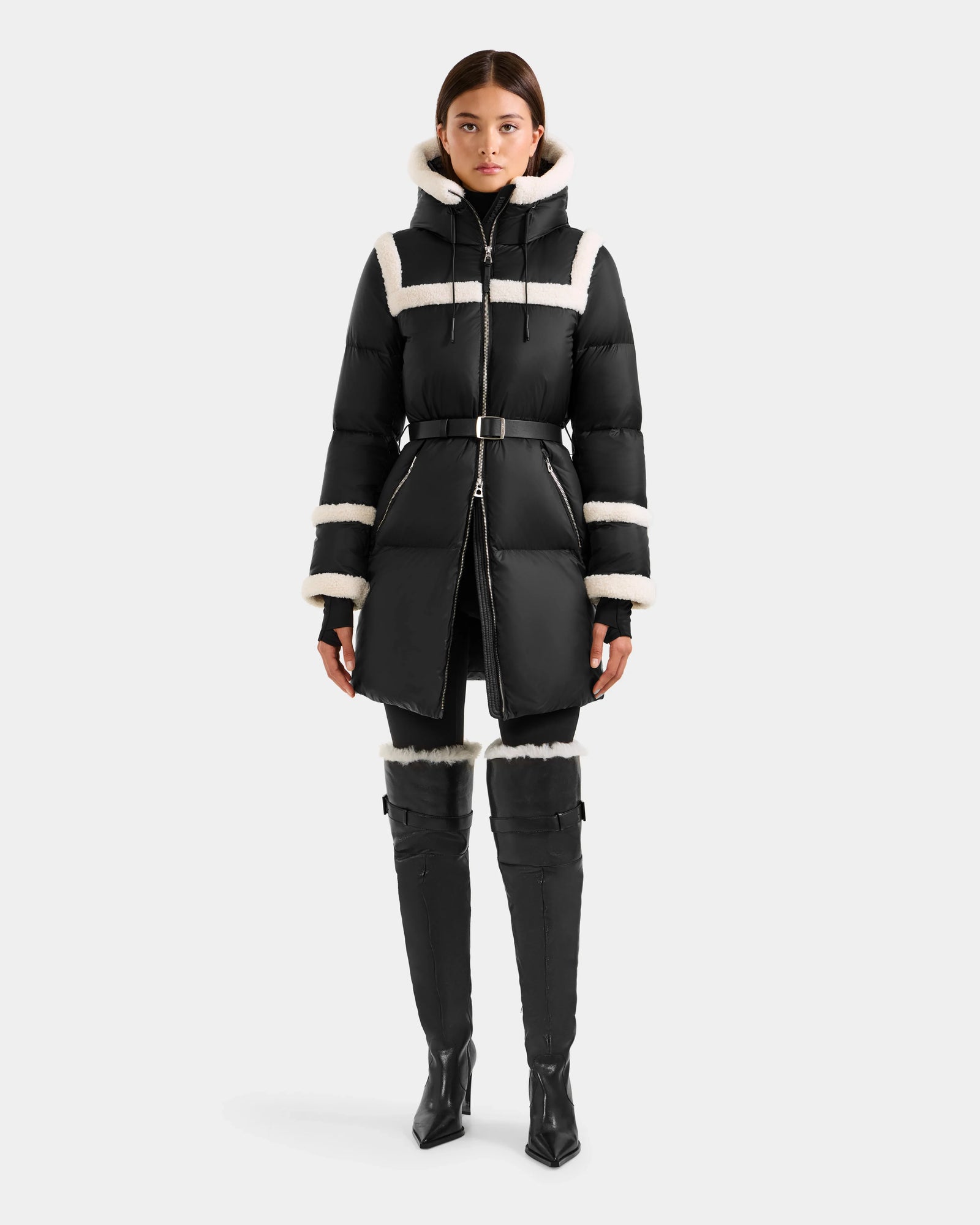 A woman faces forward in the Rudsak JULIANA L - L125535 Black/Vanilla belted puffer coat with white shearling trim, paired with black high-heeled over-the-knee boots. The background is plain white.