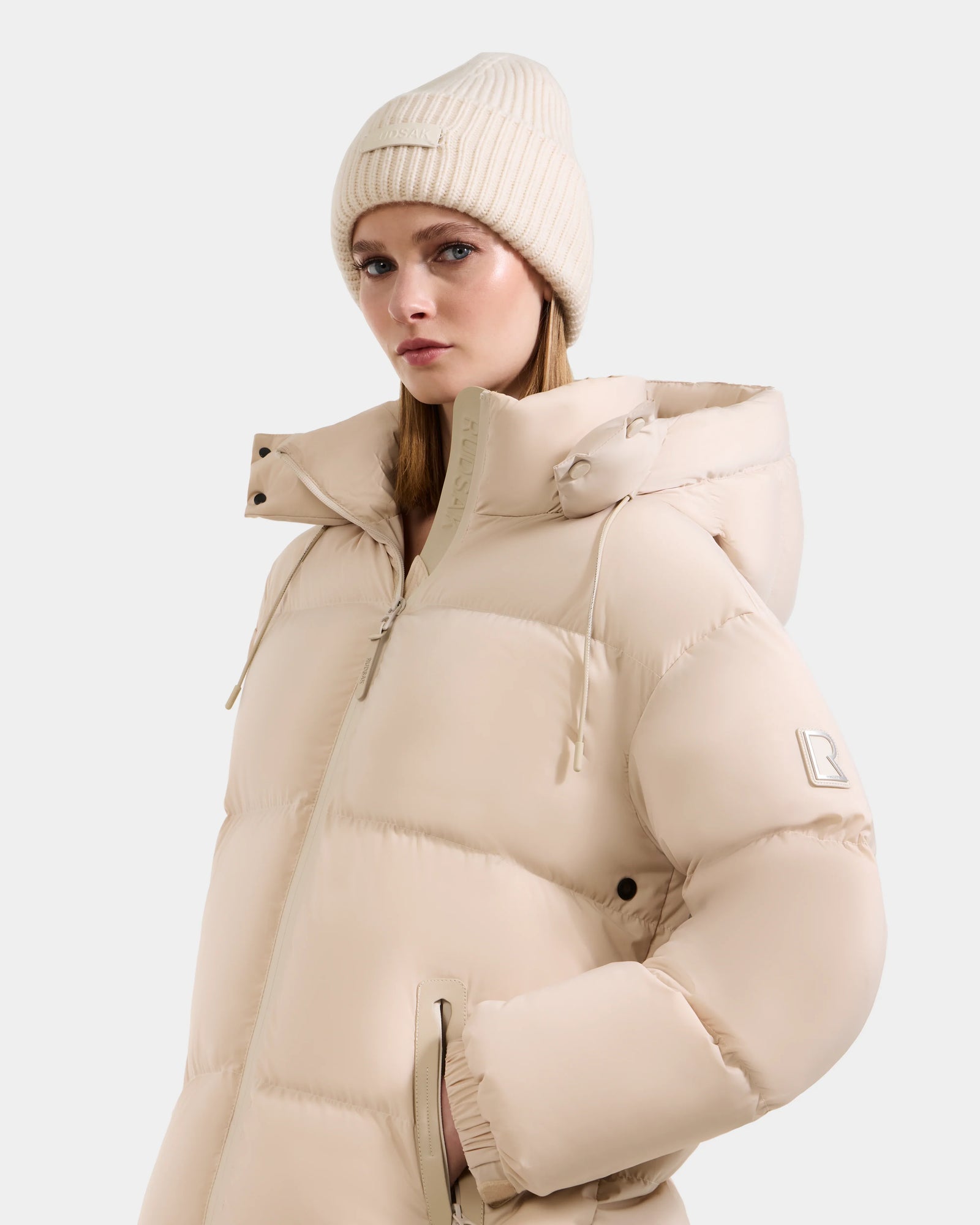 A woman wearing the Rudsak KARLEE L - 8125682 Vanilla long down puffer jacket with a hood and matching beanie stands against a plain white background, looking slightly to the side with a neutral expression.