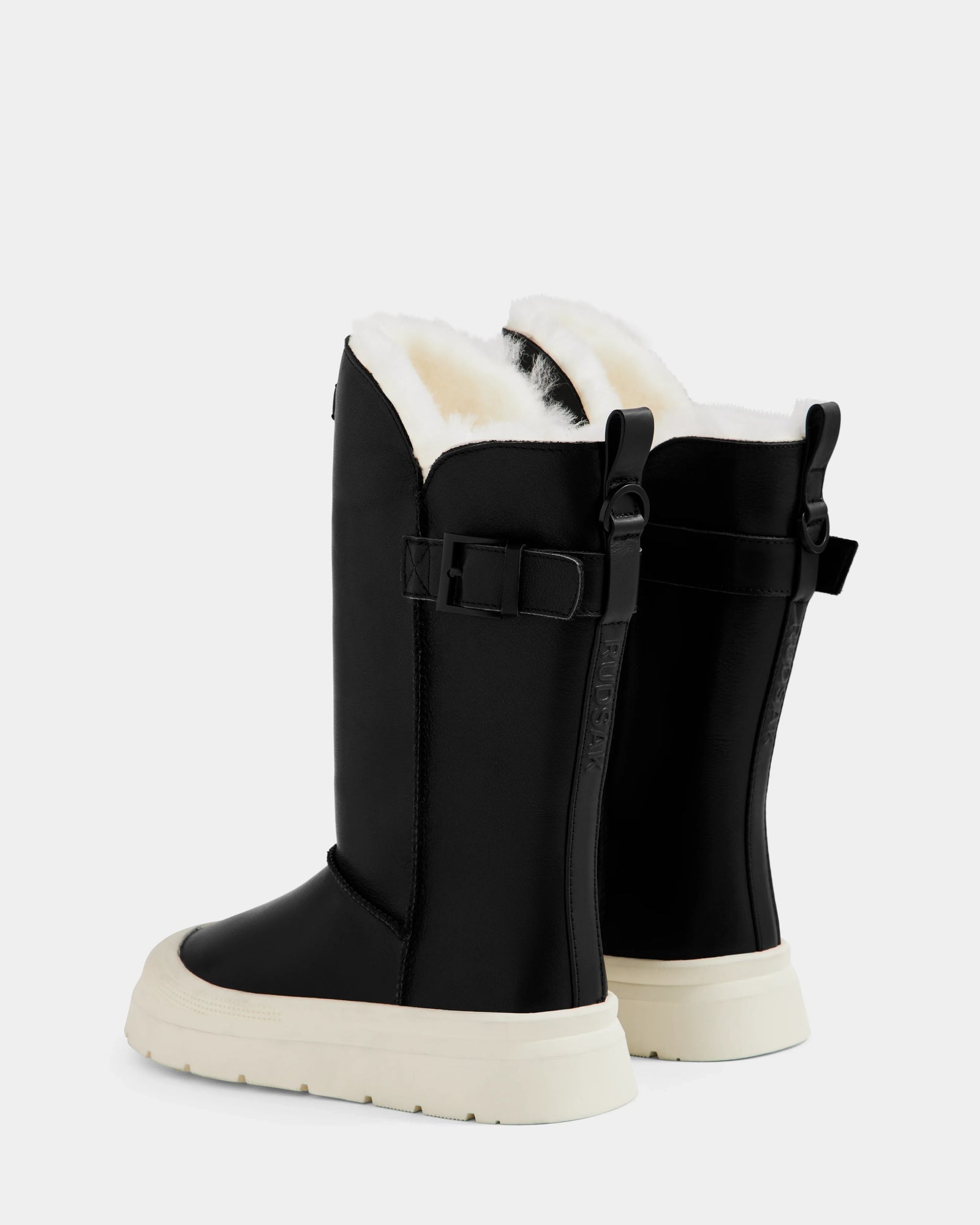 The Rudsak KLAIR L - L225519 Black women’s leather boots feature white soles, cozy fleece lining, side buckles, and a high-top design. Shown from the back and side angles against a plain light background.