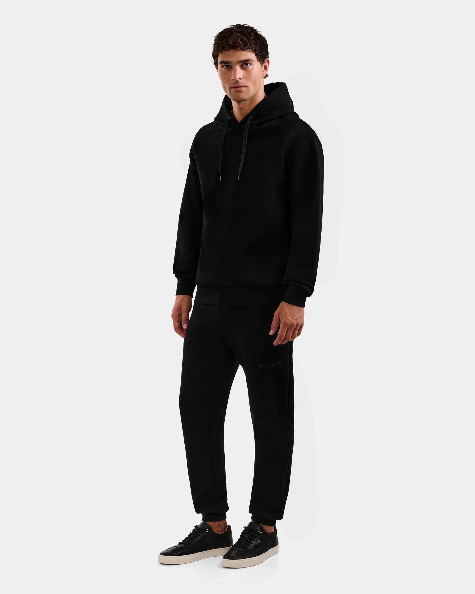 A man stands against a plain white background, wearing the Rudsak LEROY - U525509 Black M hoodie and matching black sweatpants, paired with black sneakers featuring white soles. He looks forward with a neutral expression.