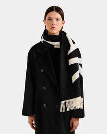 A woman wearing the Rudsak LEV - U425506 Black long coat and a reversible unisex scarf stands against a plain light background, looking at the camera. This cold-weather accessory adds style and comfort to her outfit.