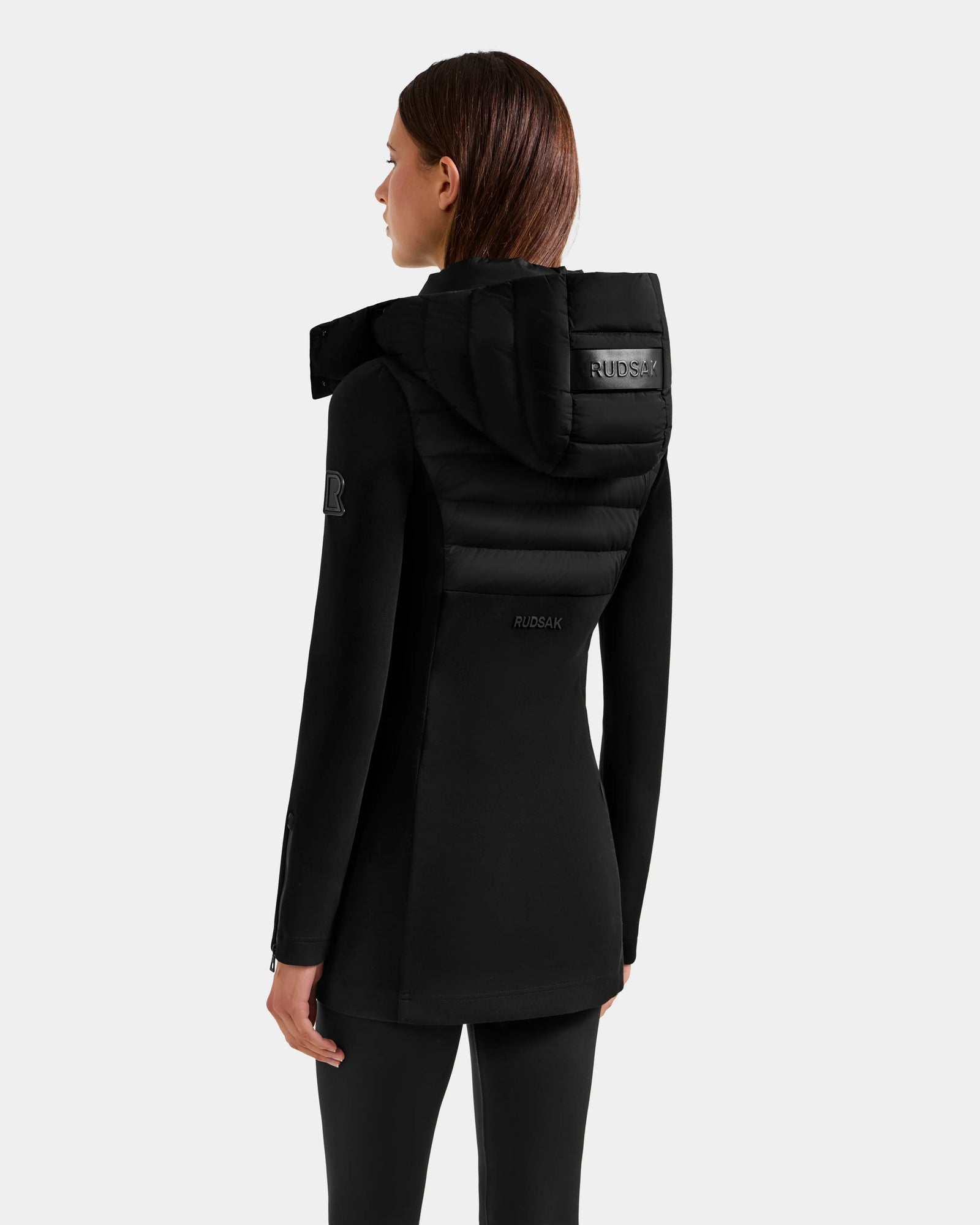A woman with straight brown hair, wearing the Rudsak MARRY M - L125519 Black mid-length hybrid coat with a padded upper back and hood, stands facing away from the camera against a plain light gray background.