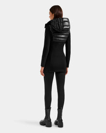 A woman faces away, wearing the Rudsak MARRY S - L125521 Black glossy shell jacket with a shiny padded hood, paired with black leggings and chunky weather-resistant boots against a plain light gray background.