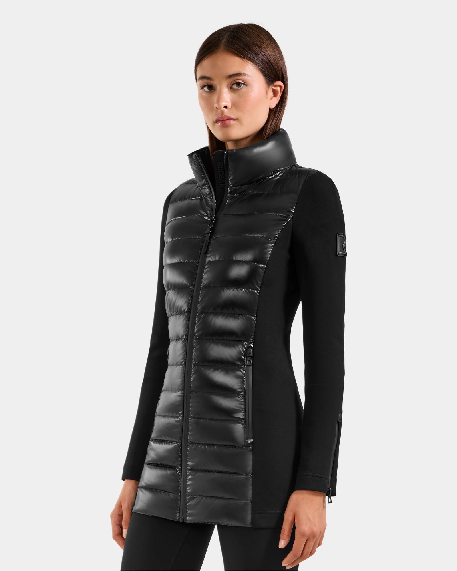 A woman wearing the Rudsak MARRY S - L125521 Black long, fitted coat with a glossy front panel and high collar stands against a plain light gray background.