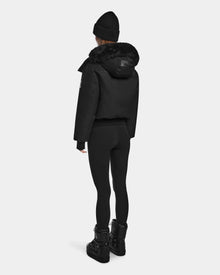 A person wearing the Rudsak MAUDE - L124522 Black/Black winter jacket, black beanie, leggings, and boots stands facing away against a plain white background.