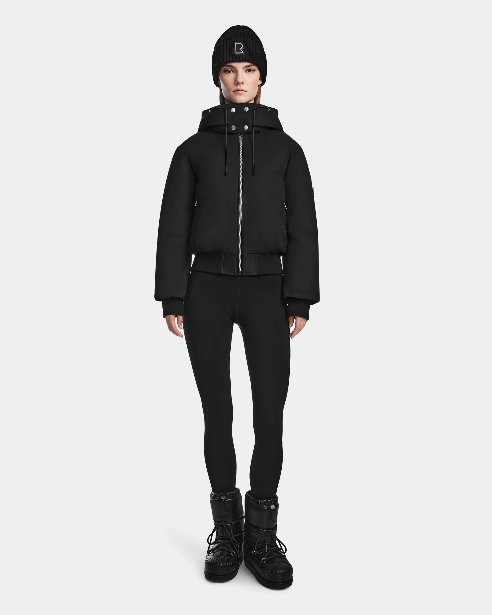 A person stands facing forward wearing the Rudsak MAUDE - L124522 Black/Black puffer jacket, a black beanie, black leggings, and winter boots against a plain light background.